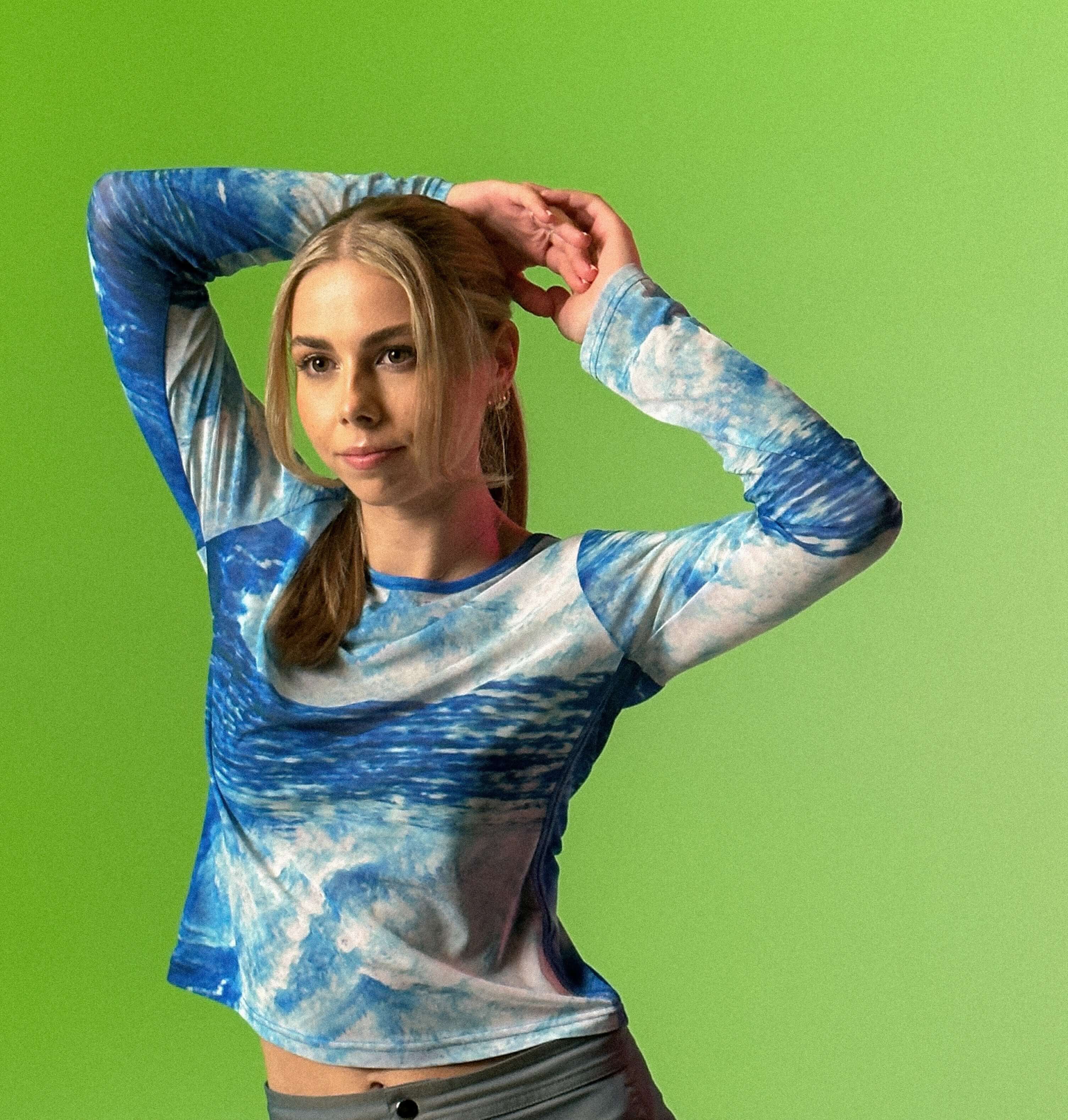 Saharaa about image posing with hands behind head in blue tie dyed beachbrains top and grey skirt with a lime green background